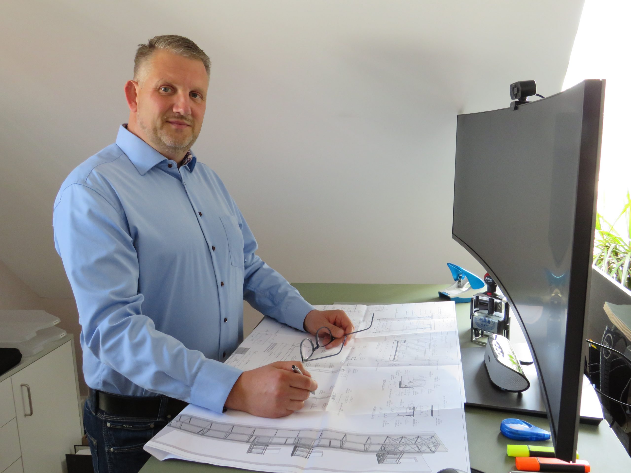 Marko Niehof steht in seinem Büro an einem Schreibtisch mit Bauplänen, trägt ein hellblaues Hemd und blickt lächelnd zur Kamera.